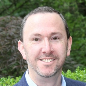 Man in blue blazer smiles in front of green foliage.