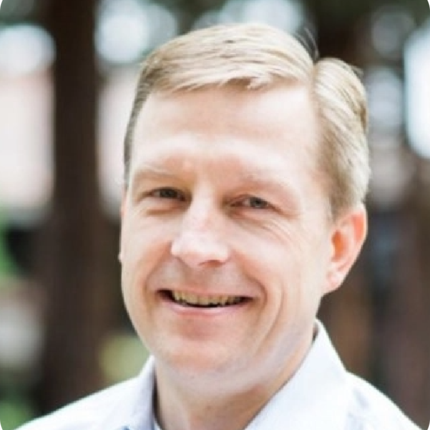 Man with blond hair smiling, wearing a light blue shirt, outdoors.