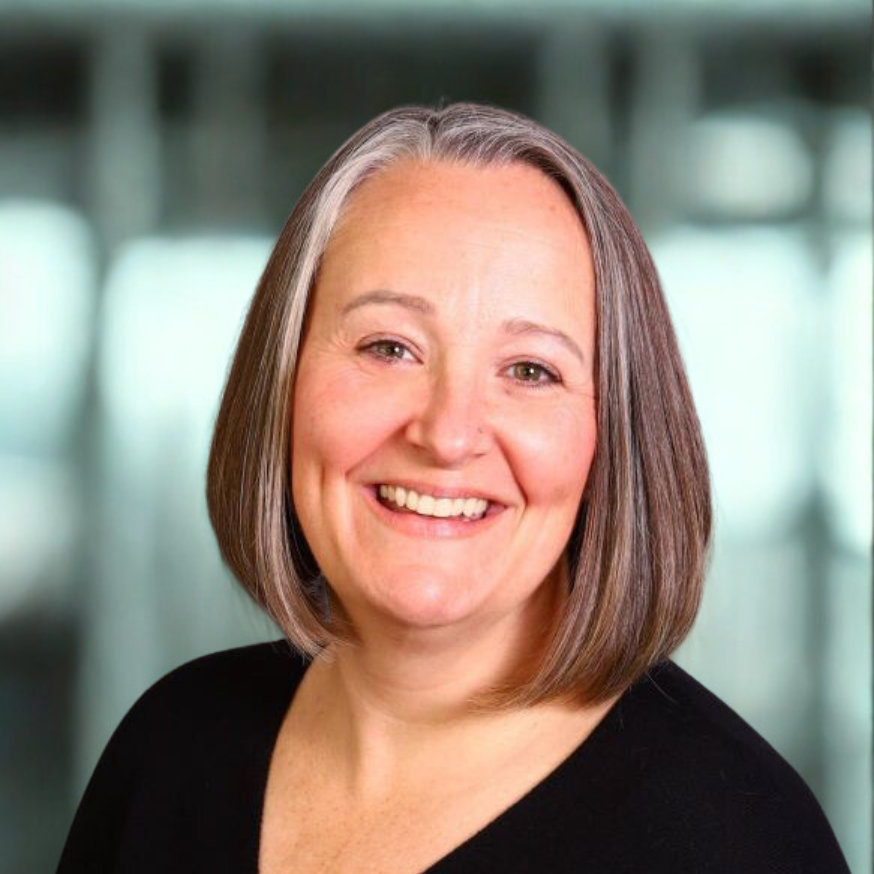 Woman with grey and brown bob smiling. Black shirt, neutral background.