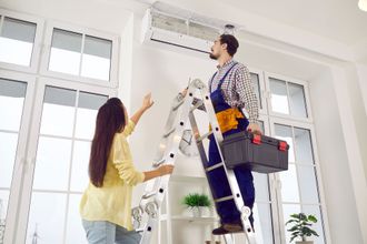 A man is standing on a ladder fixing an air conditioner while a woman looks on.