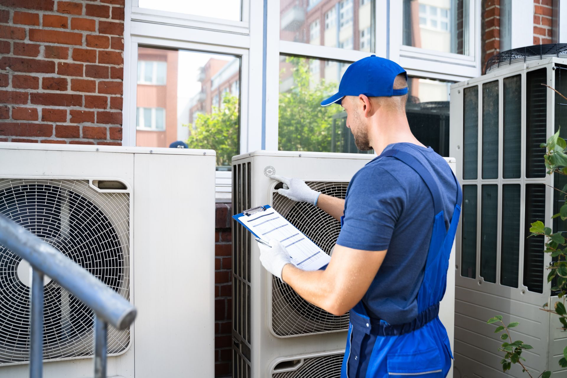 A man is working on an air conditioner outside of a building while holding a clipboard.