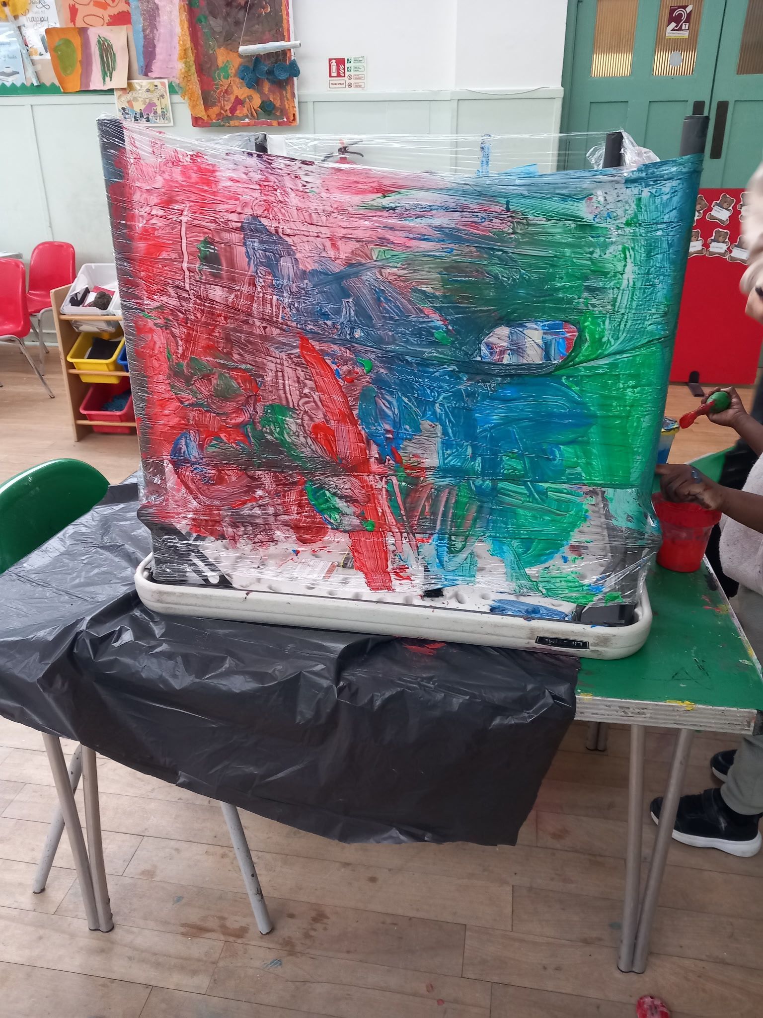 A child painting with red, blue, and green on a plastic-wrapped object in a classroom.