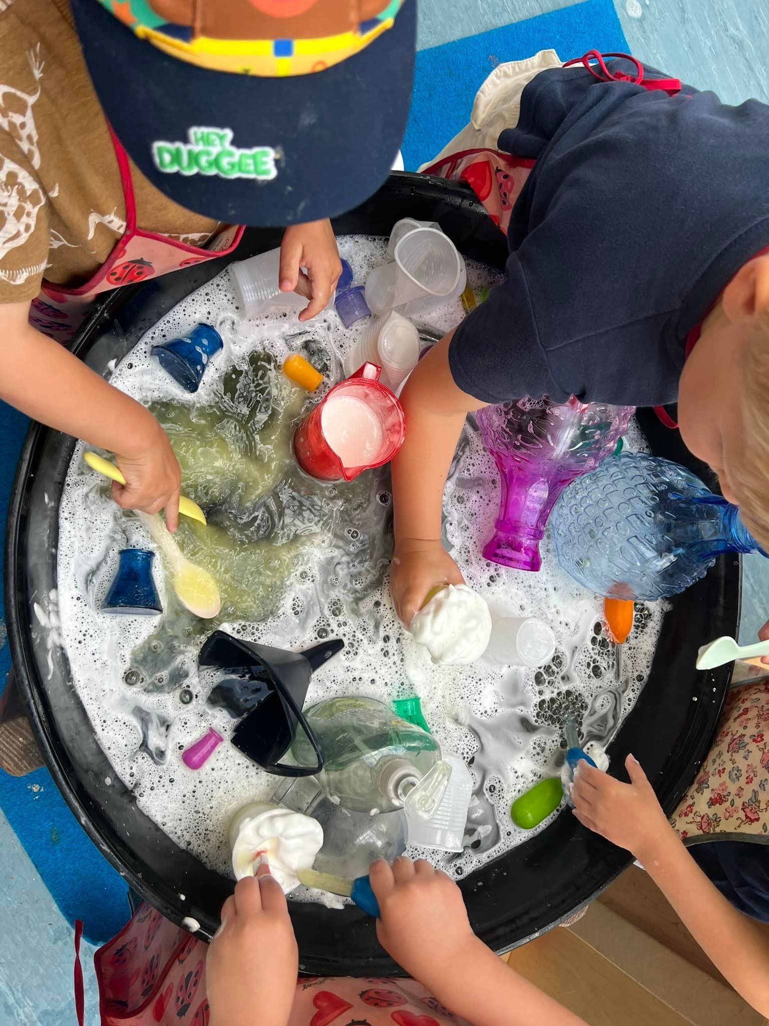 Children playing with soapy water in a black tub, using cups and spoons.