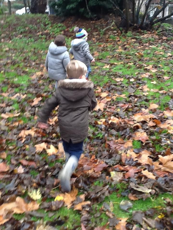 Three children run through autumn leaves on a grassy hill, wearing coats and hats.