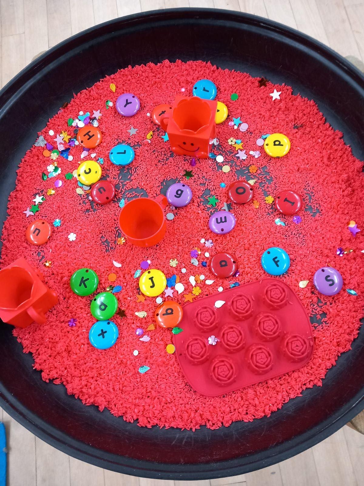 Sensory play tray with red sand, colorful letters, and toys.