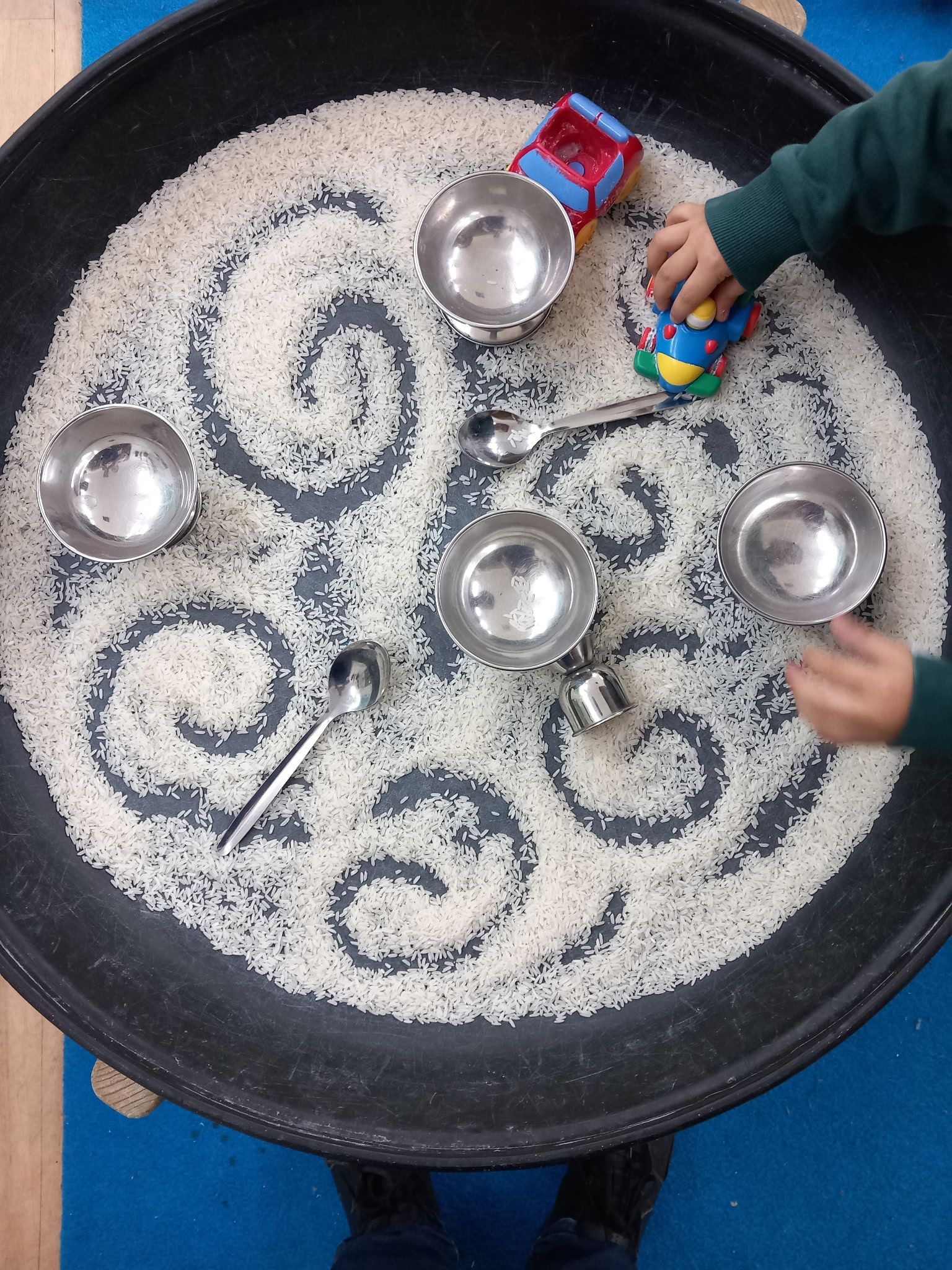 Child playing with toys and metal bowls in a tray of rice; hands visible.