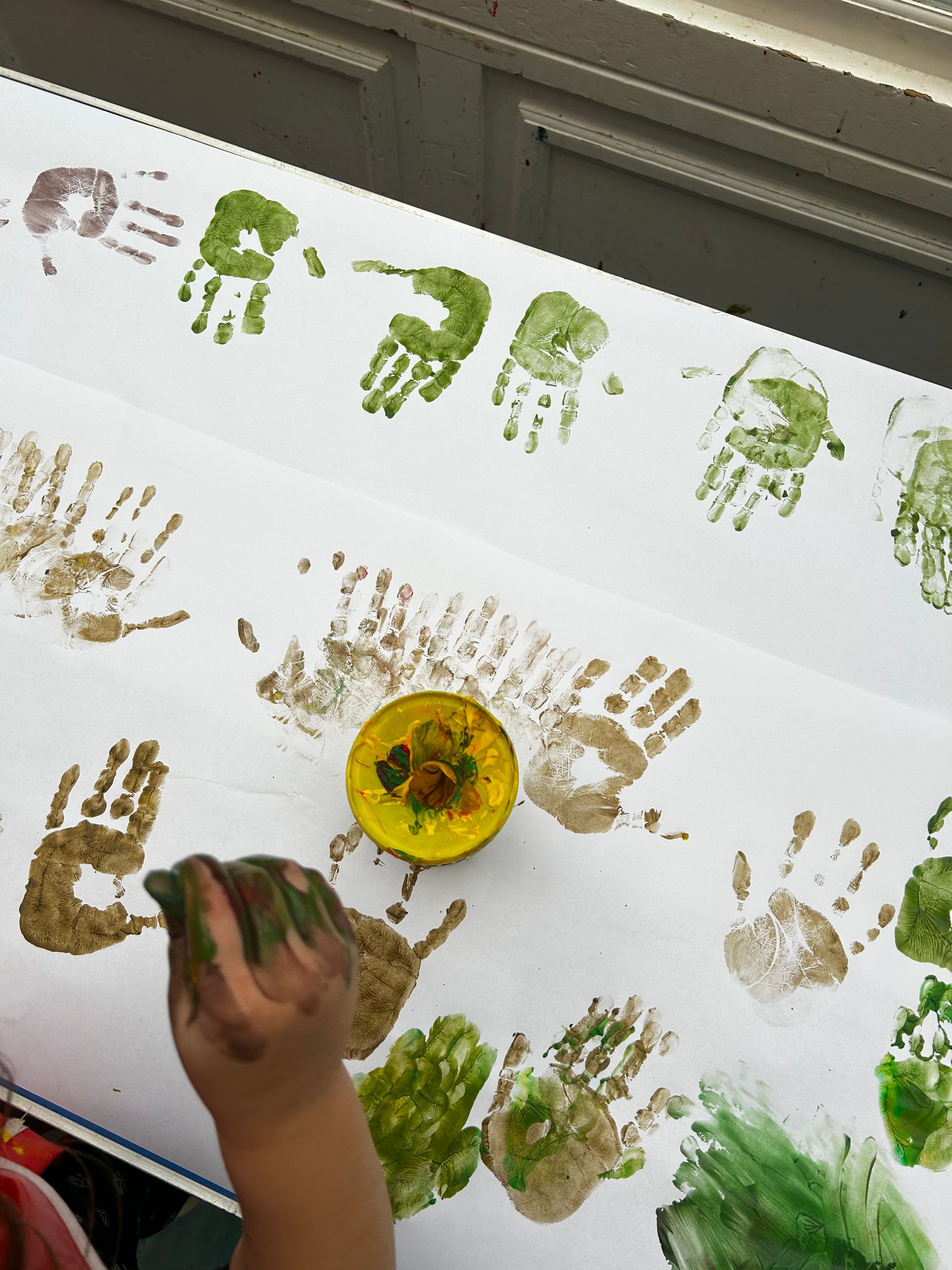 Child hand painting handprints on white surface with green and brown paint.