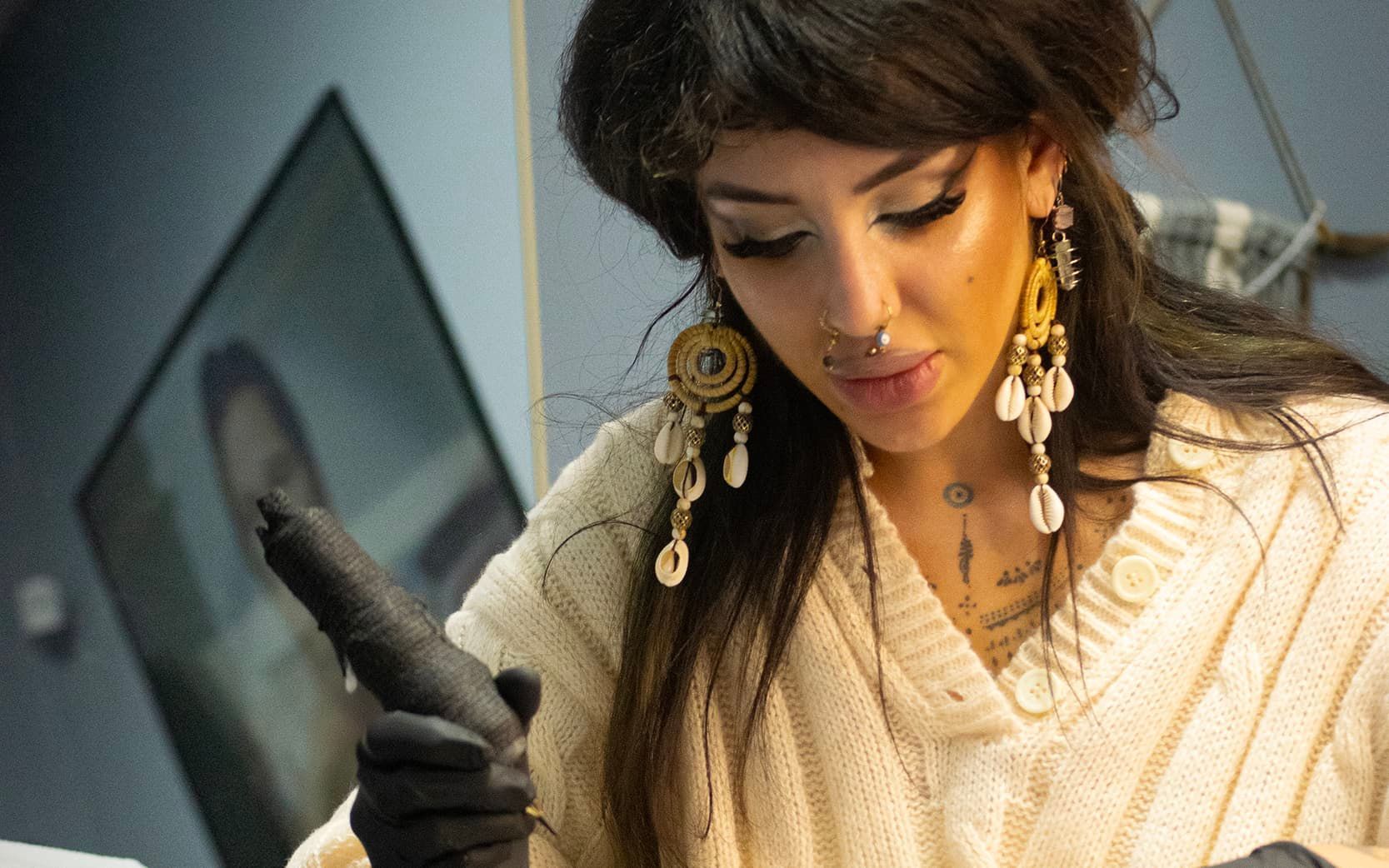 Woman in cream sweater and black gloves examining a small object indoors, wearing gold earrings.