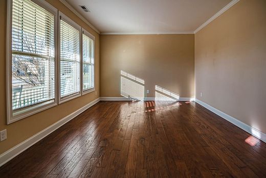 An empty room with dark wood flooring, tan walls, and three windows letting in sunlight, creating striped patterns.