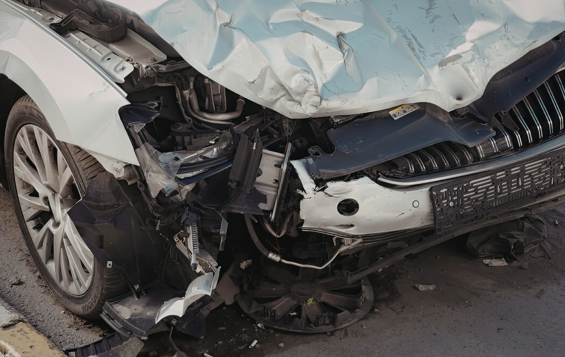 Close-up of the heavily damaged front end of a white car, showing a crushed hood, bumper, and shattered headlight.