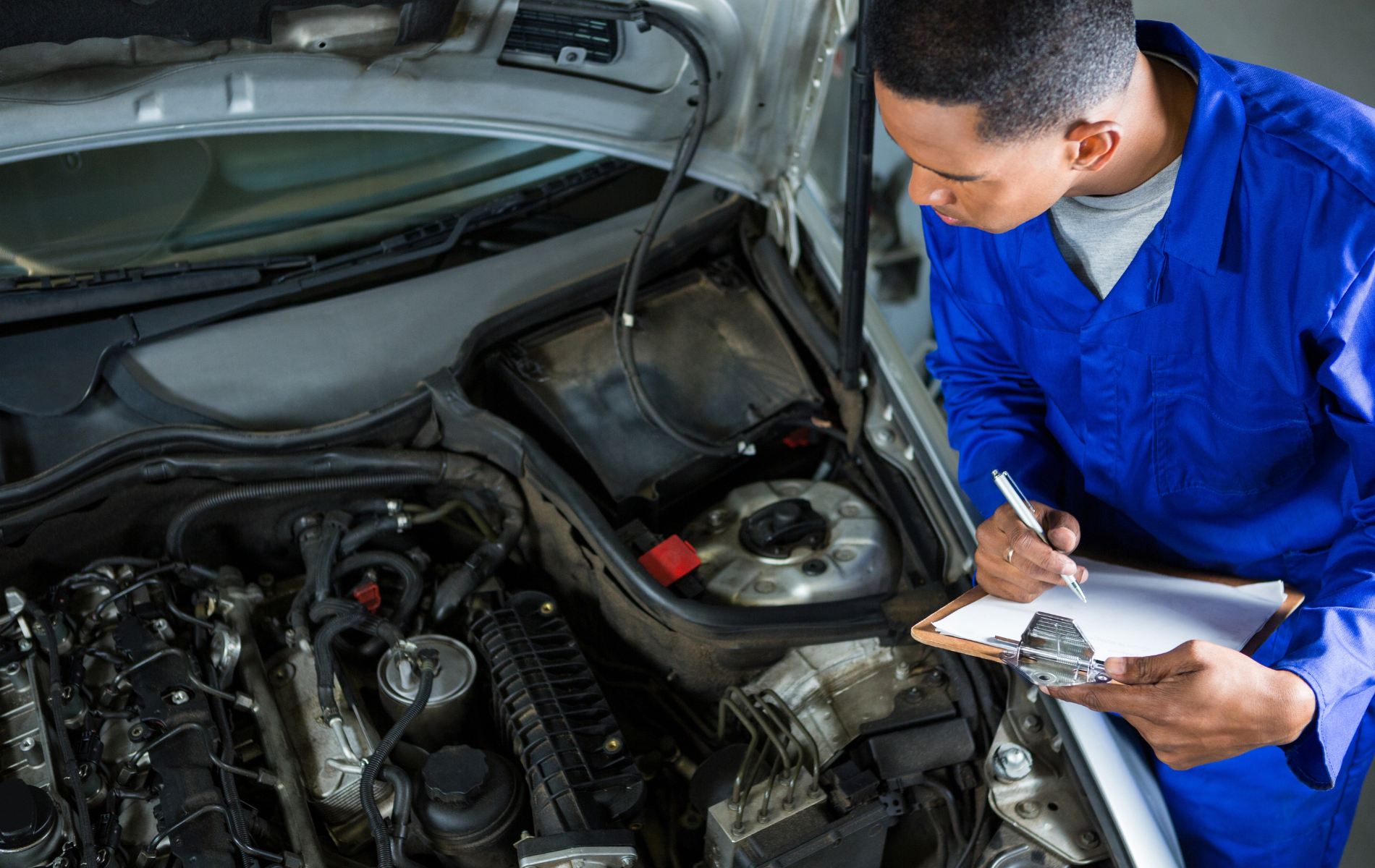 A mechanic in a blue uniform inspecting a car engine while taking notes on a clipboard.