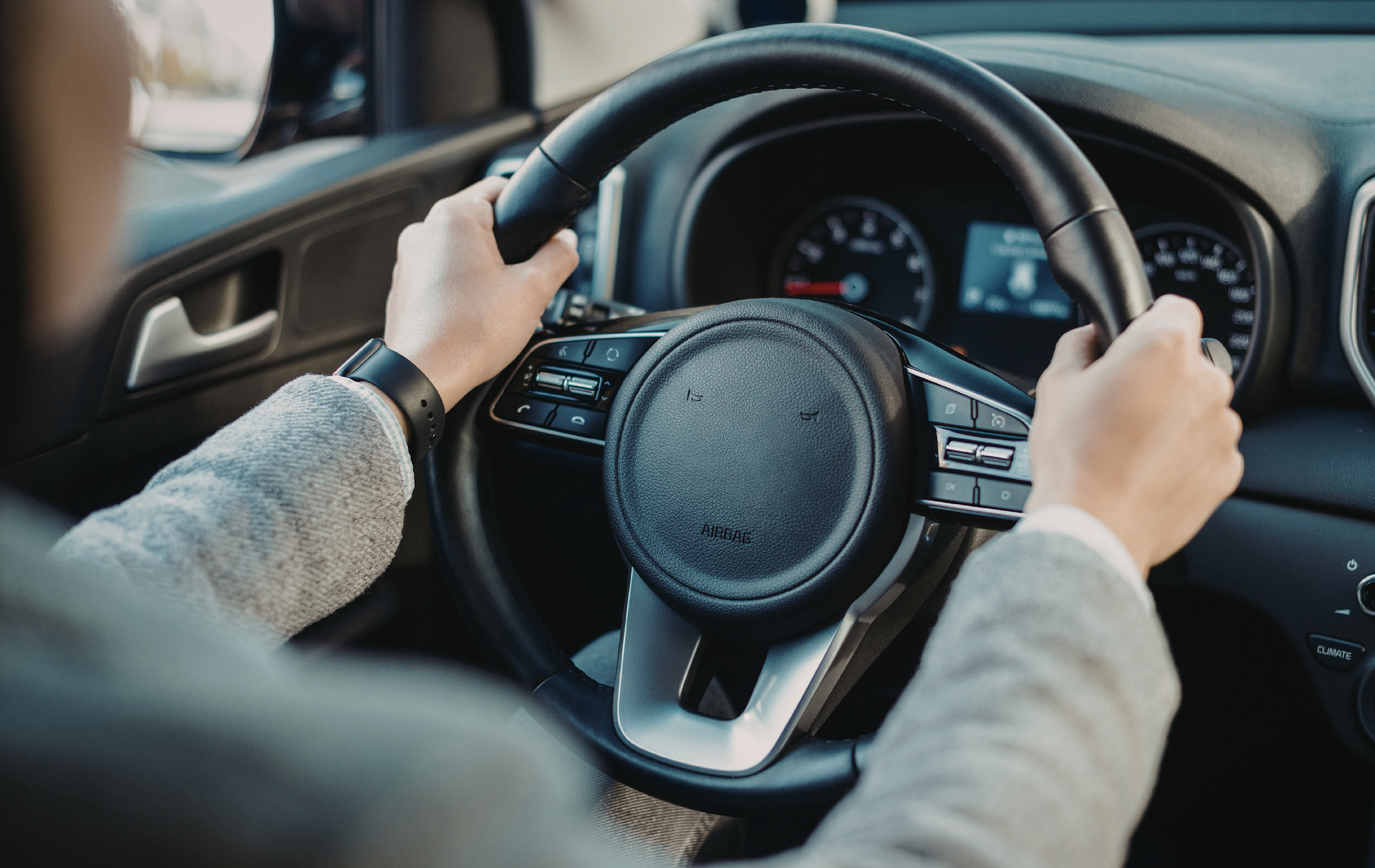 A person’s hands grip the steering wheel of a car from the driver's perspective, with the dashboard visible ahead.
