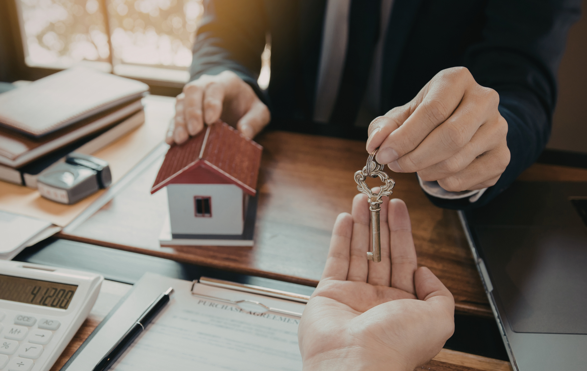 A person in a suit hands a set of keys to another person, with a small model house on a desk beside them.