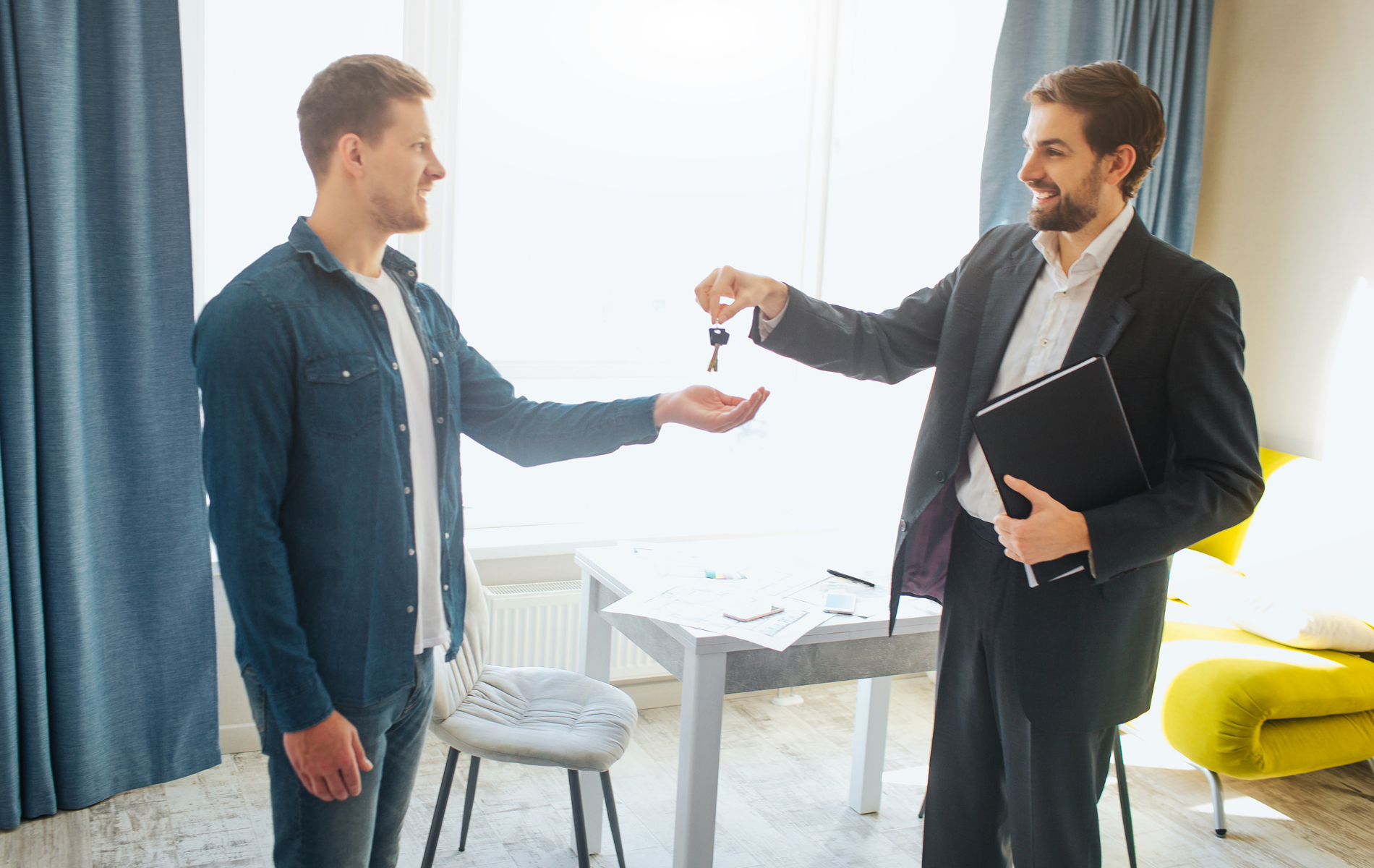 A business professional in a suit hands a set of keys to a person in a blue shirt inside a bright, modern room.