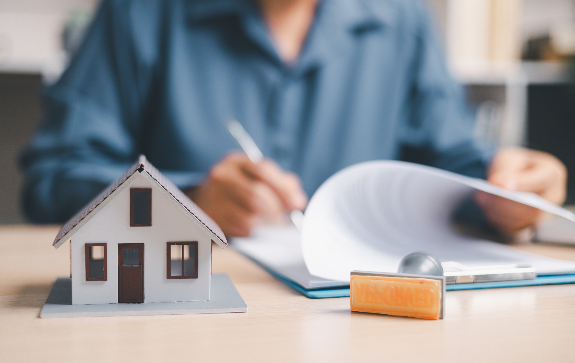 A model house and a rubber stamp sit on a desk as a person reviews documents in the background.