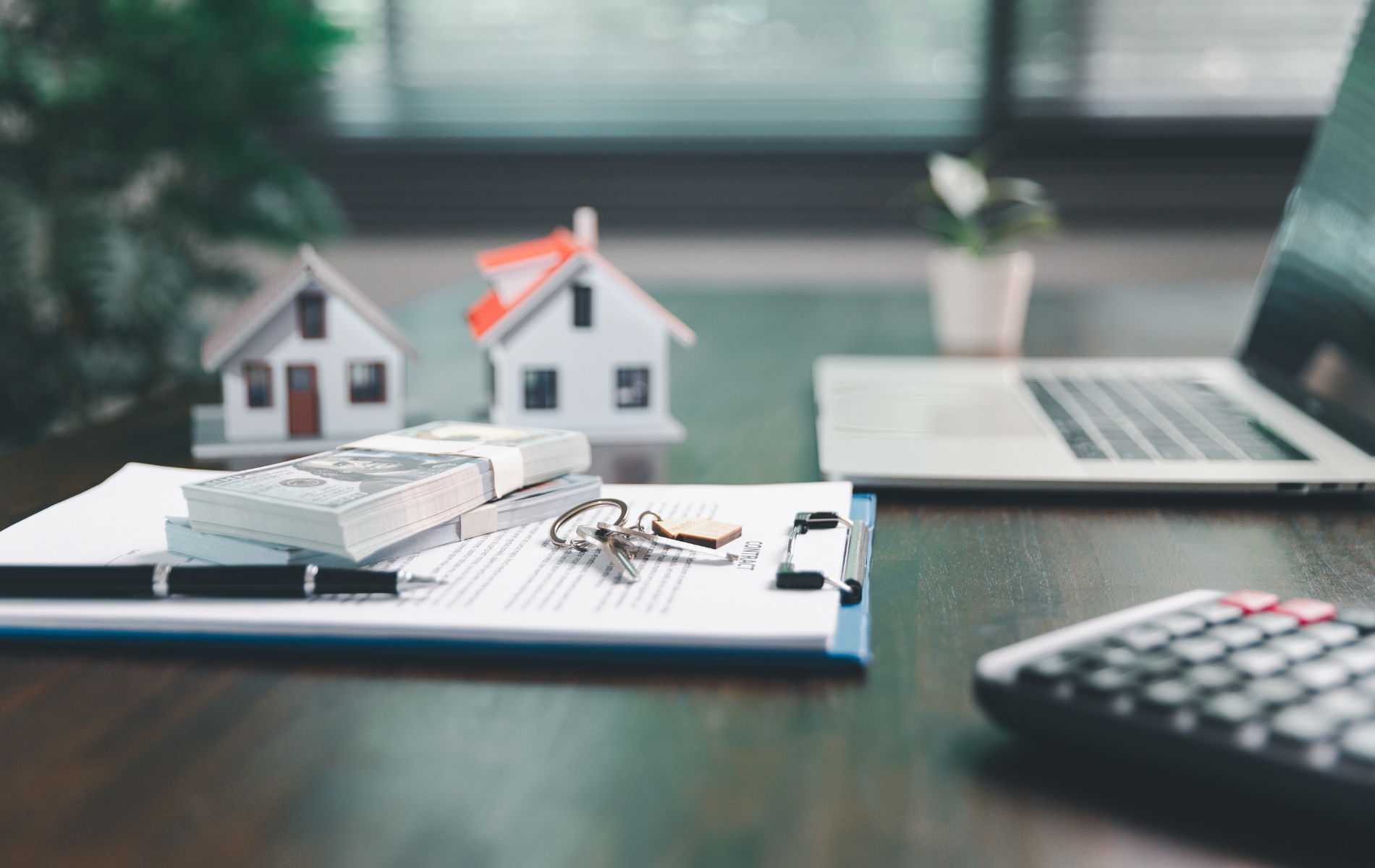 A stack of cash, keys, and model houses rest on a document near a laptop and calculator on a dark office desk.