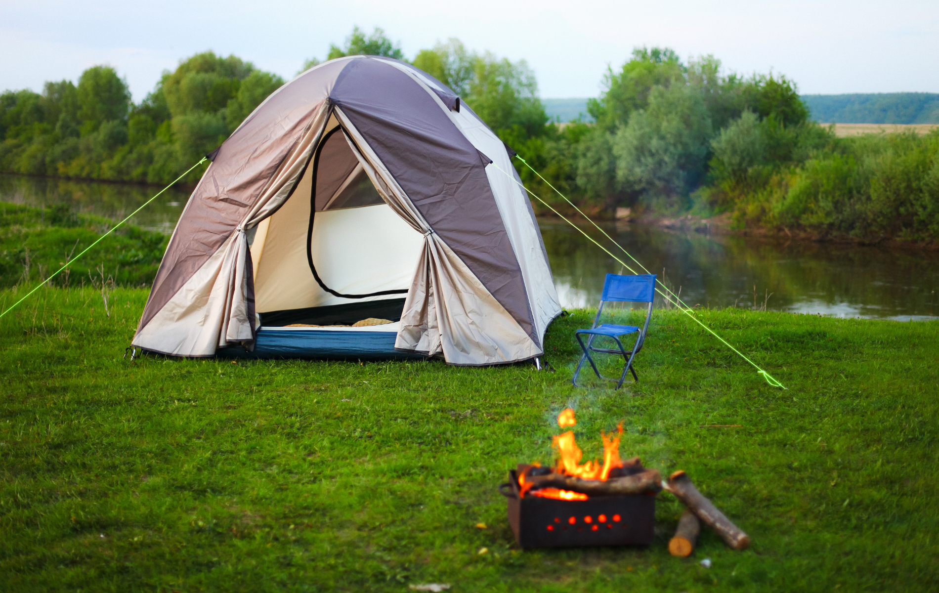 Camping scene: tent by river, campfire burning, folding chair on grass.