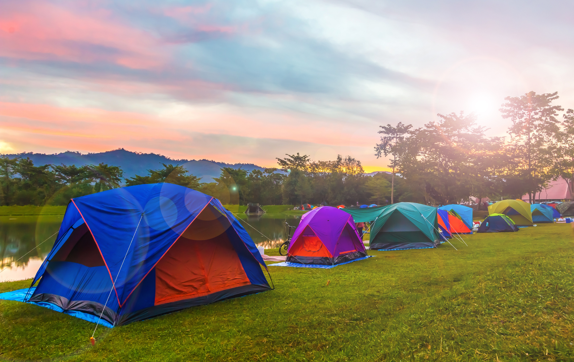 Colorful tents arranged on a grassy field beside a lake during a sunset with rolling hills in the background.