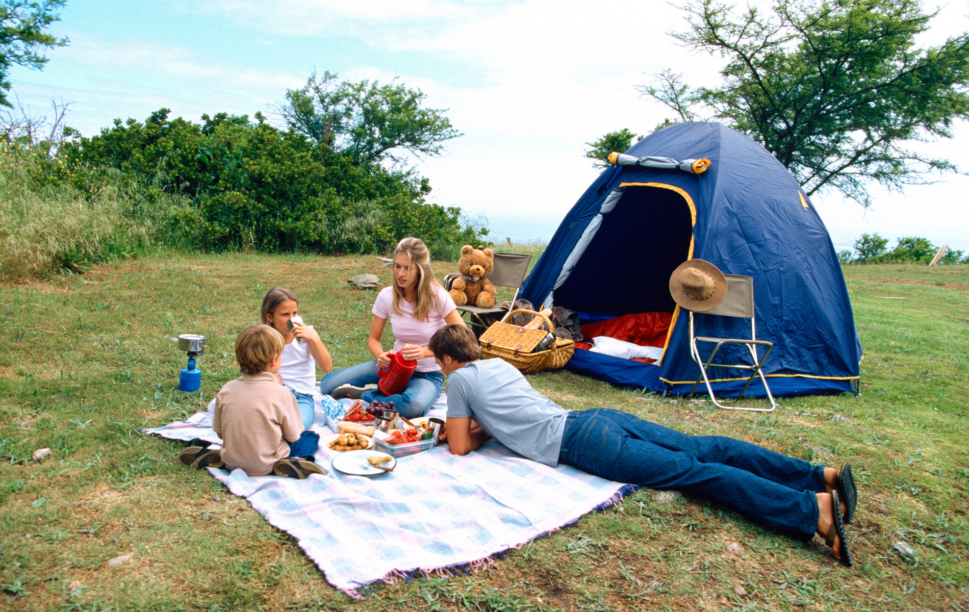 A family sits on a blanket during a picnic in a grassy field next to a blue tent.
