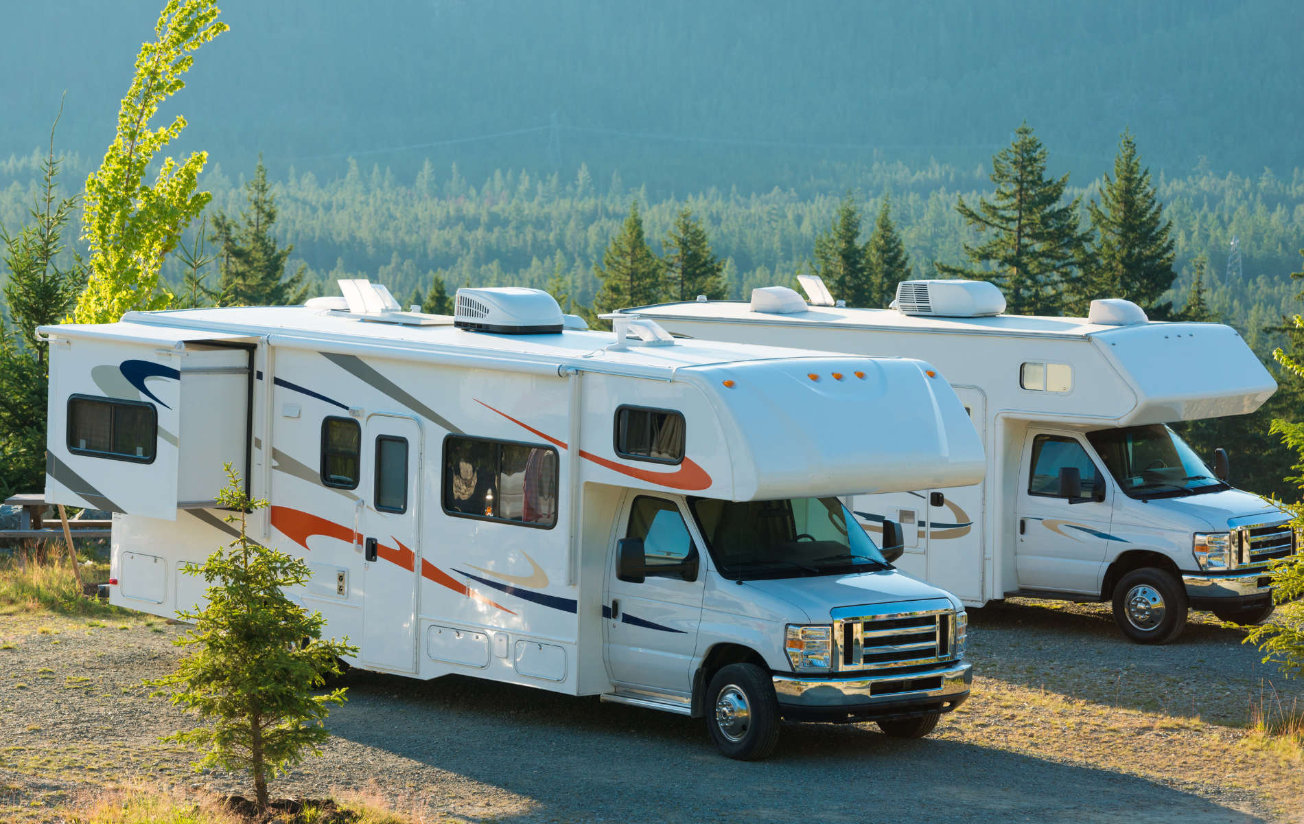 Two white RVs parked outdoors with mountain and tree background.