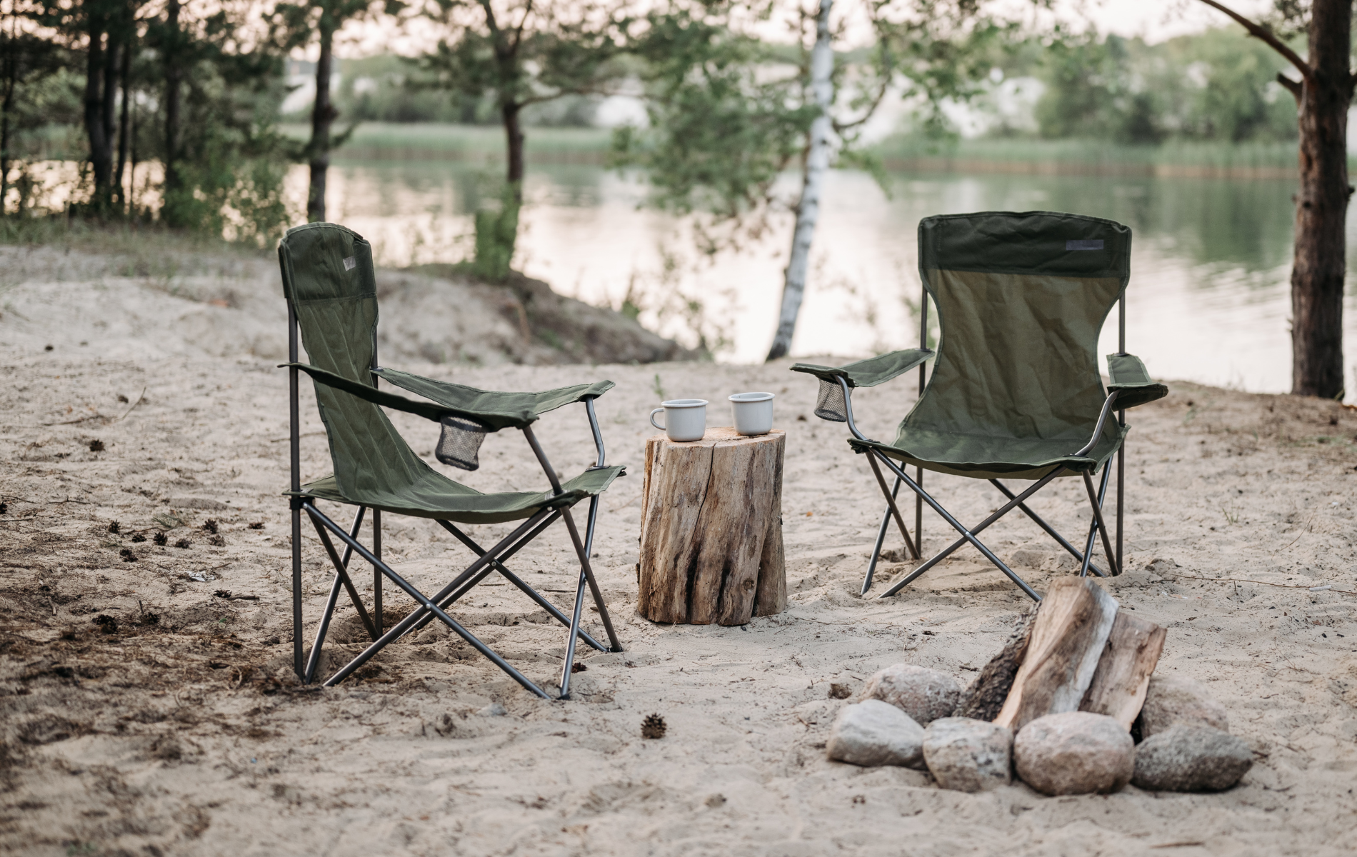 Two green camping chairs face a lake, with a wooden stump table and campfire on the sand.