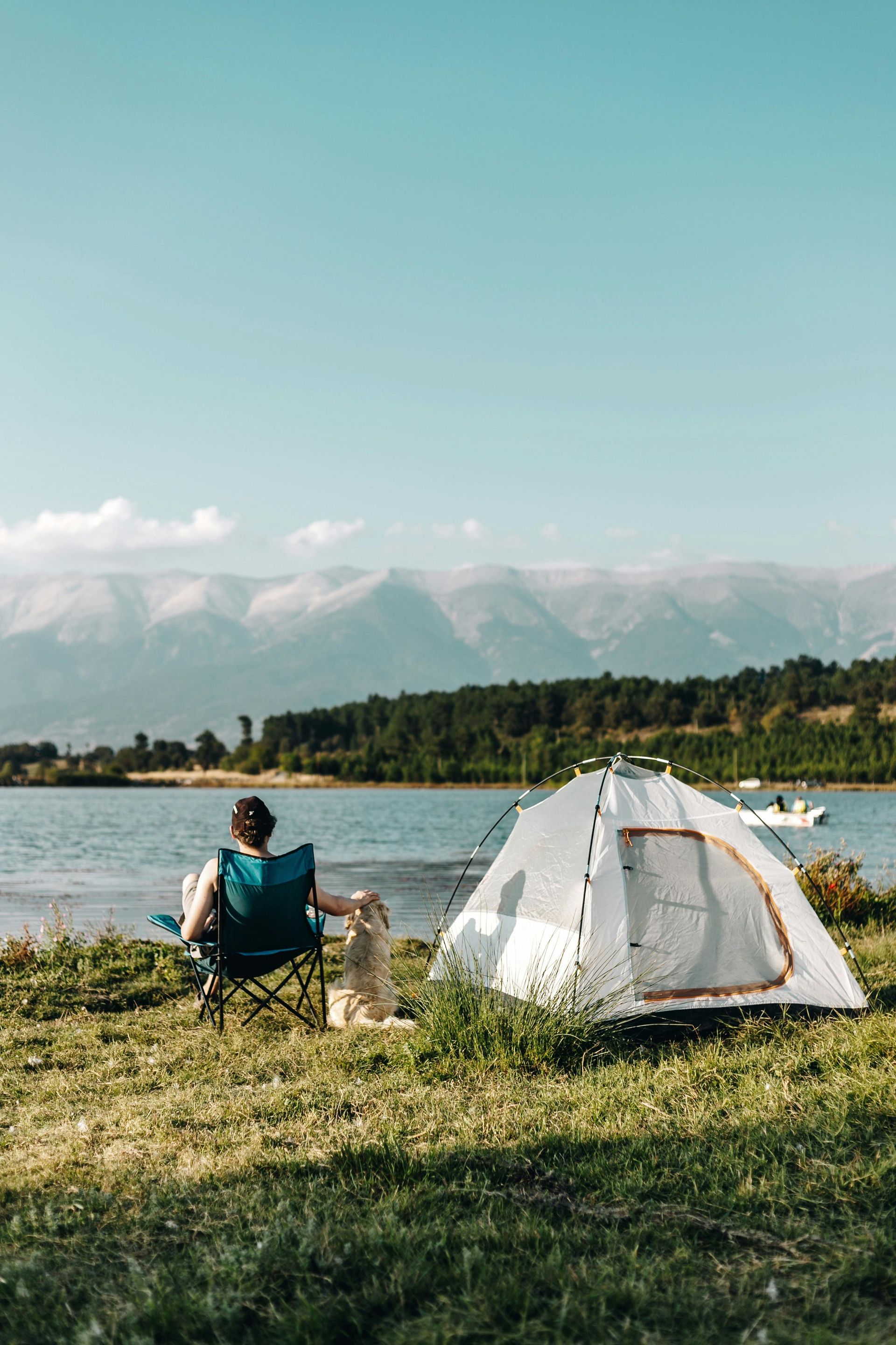 A person sits in a camping chair by a lake next to a white tent, with mountains and forest in the distance.