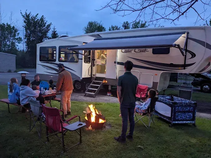 Family around campfire, RV in background, awning extended. People sitting at table, one standing. Dusk setting.