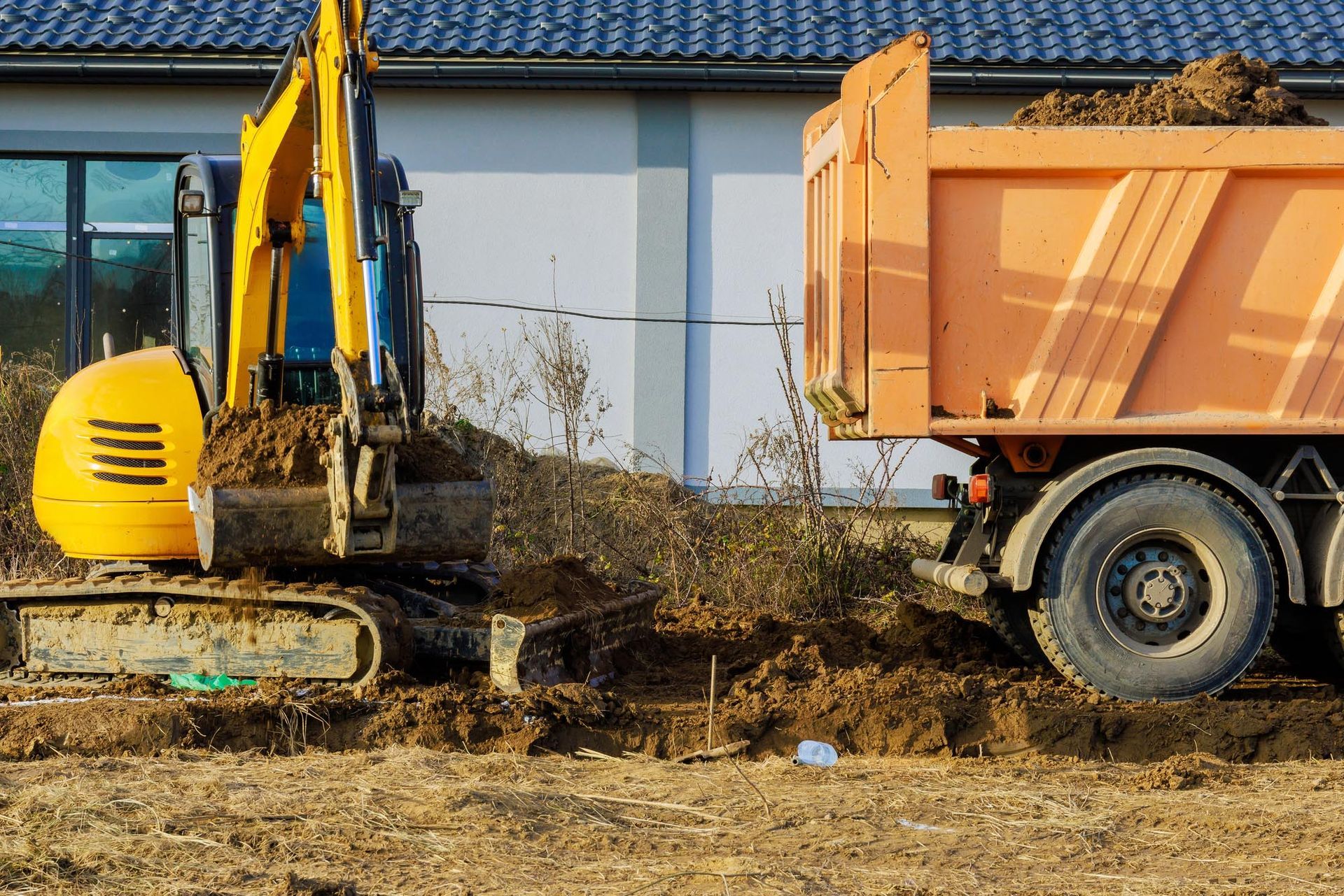 In un cantiere edile di fronte a un edificio, un escavatore giallo scava del terreno accanto a un autocarro ribaltabile arancione.
