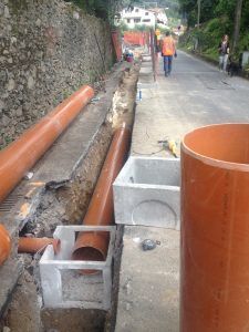 Cantiere stradale con tubi fognari in plastica arancione e pozzetti di drenaggio in cemento in uno scavo a cielo aperto.