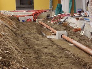 Cantiere edile con un tubo in PVC marrone, sorretto da blocchi di cemento, che attraversa uno scavo di terra vicino a un muro giallo.