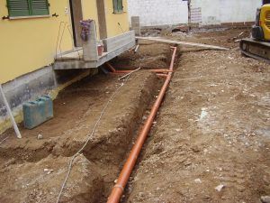 Tubi di drenaggio in PVC arancione installati in una trincea a lato della facciata esterna di un edificio residenziale giallo in costruzione.