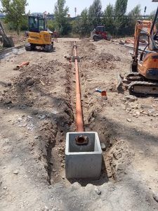 In un cantiere si può notare un lungo tubo arancione posato in una trincea, che conduce a una vasca quadrata in cemento, dove sono presenti due escavatori.