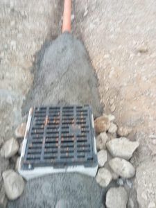 Una griglia di drenaggio nera installata su una base di cemento, collegata a un tubo arancione incastonato in uno strato di ghiaia.