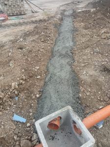 In un cantiere edile, una trincea di drenaggio riempita di cemento che conduce a una vasca di raccolta prefabbricata grigia con tubi di drenaggio arancioni.