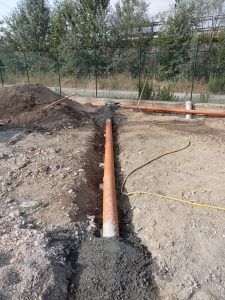 Un tubo di drenaggio arancione posato in una trincea circondata da terra, con cemento fresco colato attorno ai giunti del tubo.