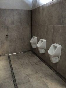 Tre orinatoi bianchi a parete in una stanza con pareti rivestite di piastrelle grigie e una canalina di scolo a pavimento lungo la parete.