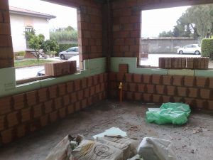 Interno di un cantiere edile con pareti in mattoni, due aperture per finestre e pannelli isolanti verdi.