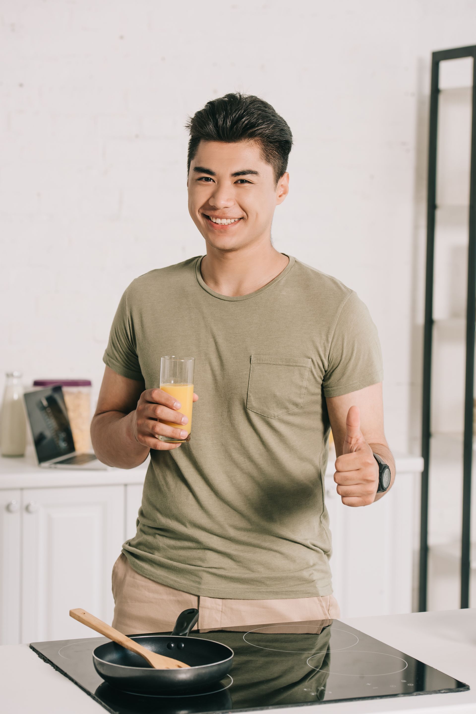 A man is standing in a kitchen holding a glass of orange juice and giving a thumbs up.