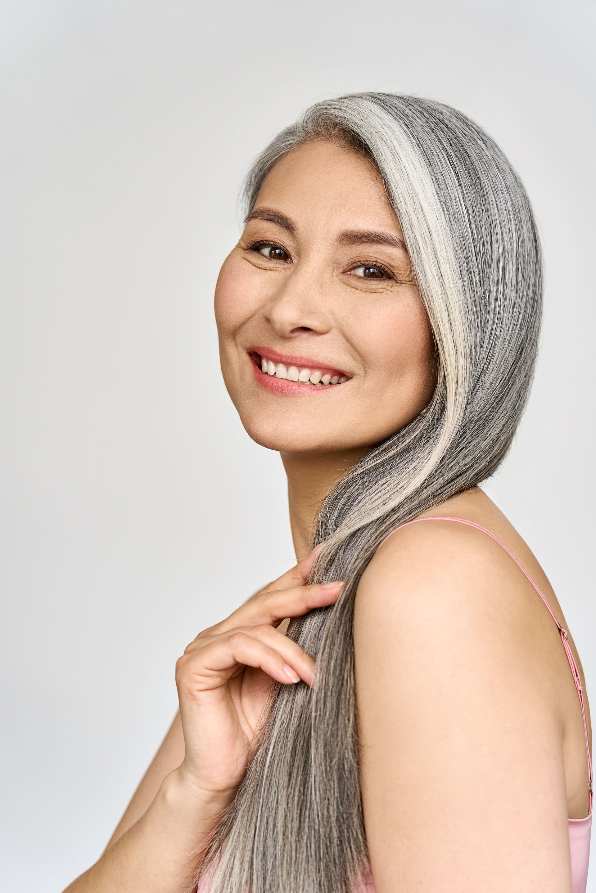 A woman with long gray hair is smiling and holding her hair.