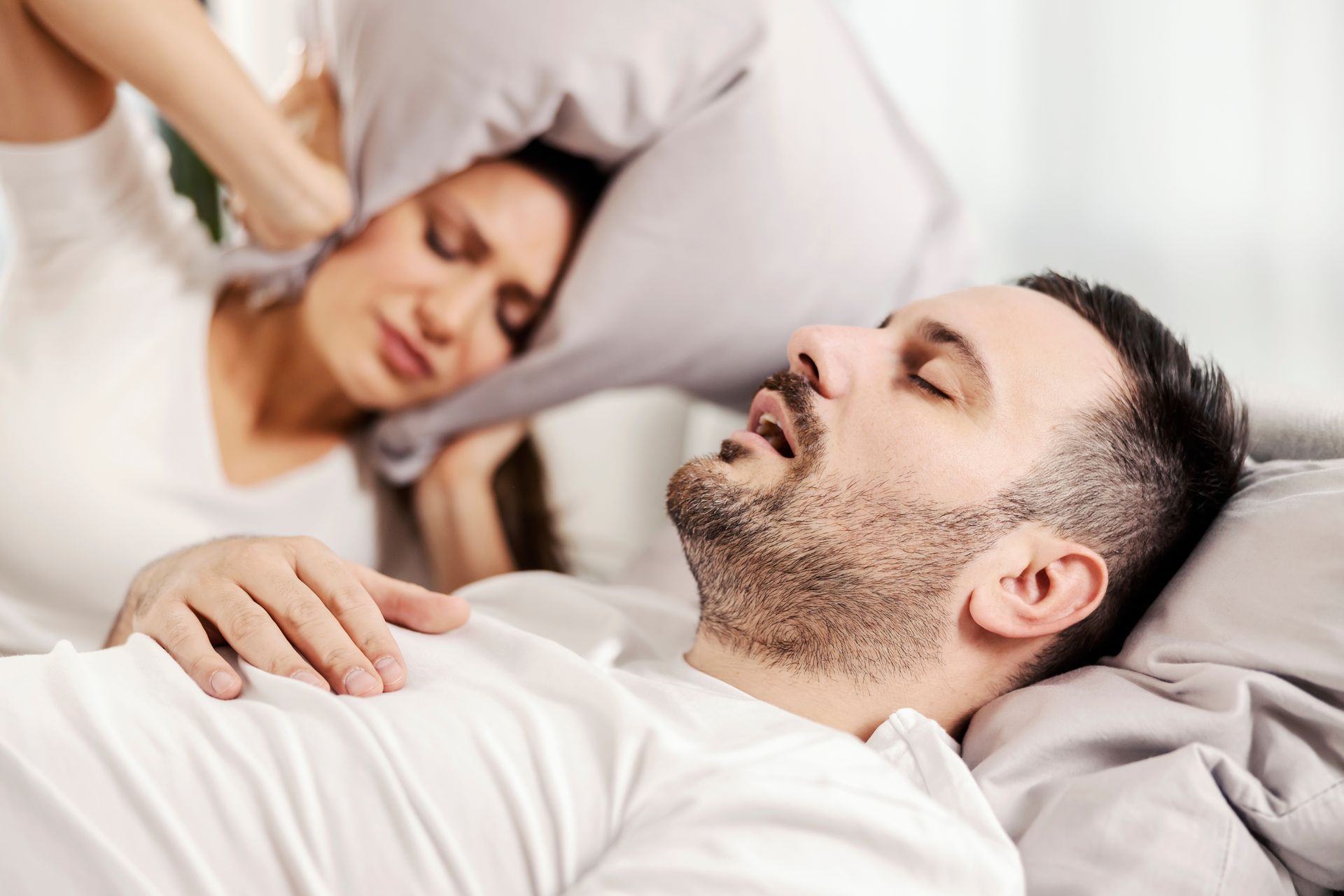 A man is snoring while a woman covers her ears with a pillow.