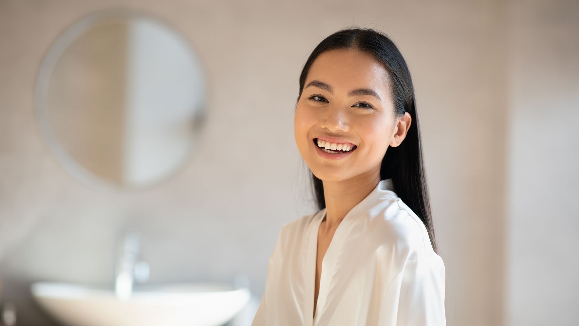 A woman in a white robe is smiling in a bathroom.