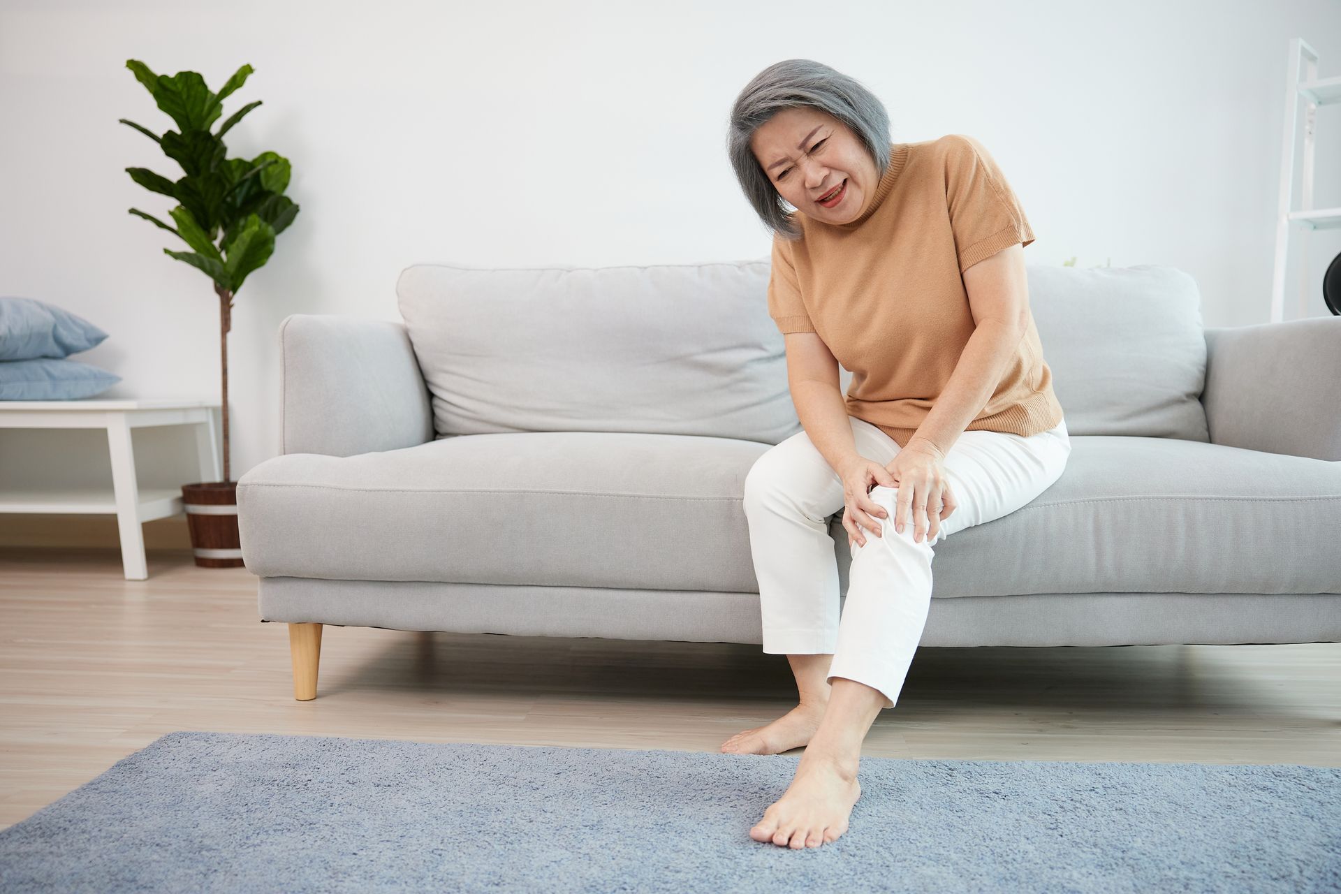 An elderly woman is sitting on a couch holding her knee in pain.