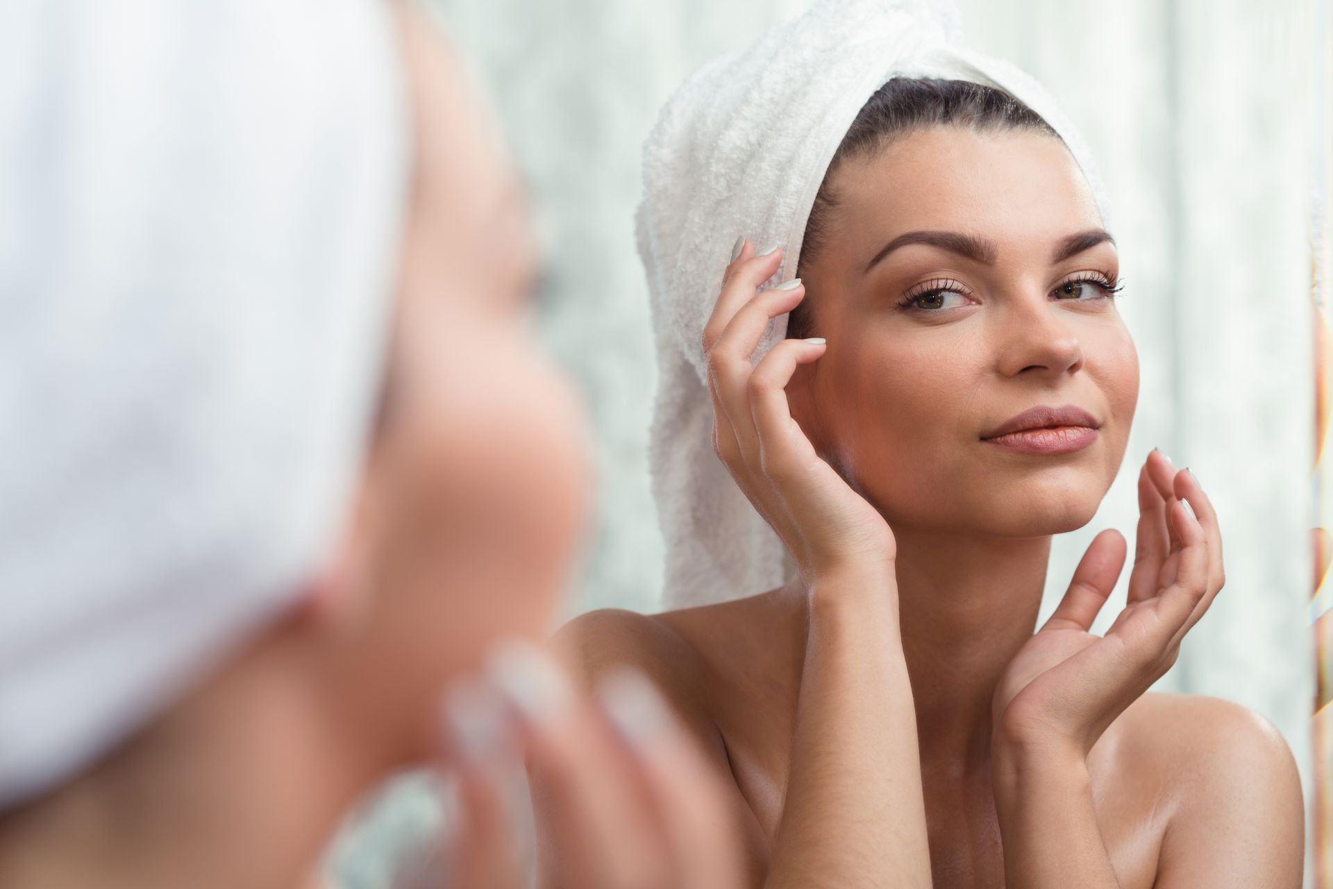 A woman with a towel wrapped around her head is looking at her face in the mirror.