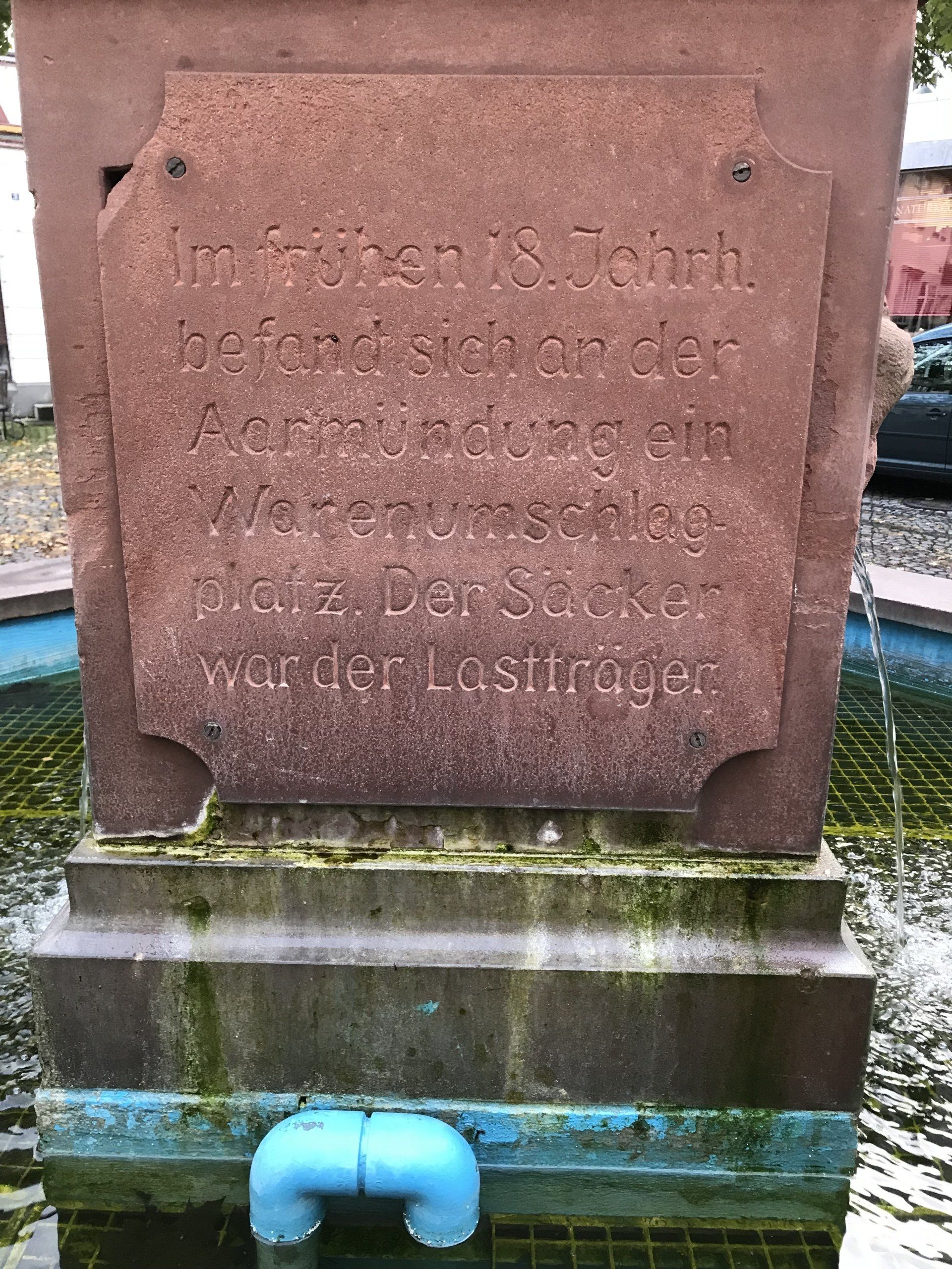 Ein Brunnen mit einer Gedenktafel darauf und einem blauen Rohr, das aus ihm herauskommt.