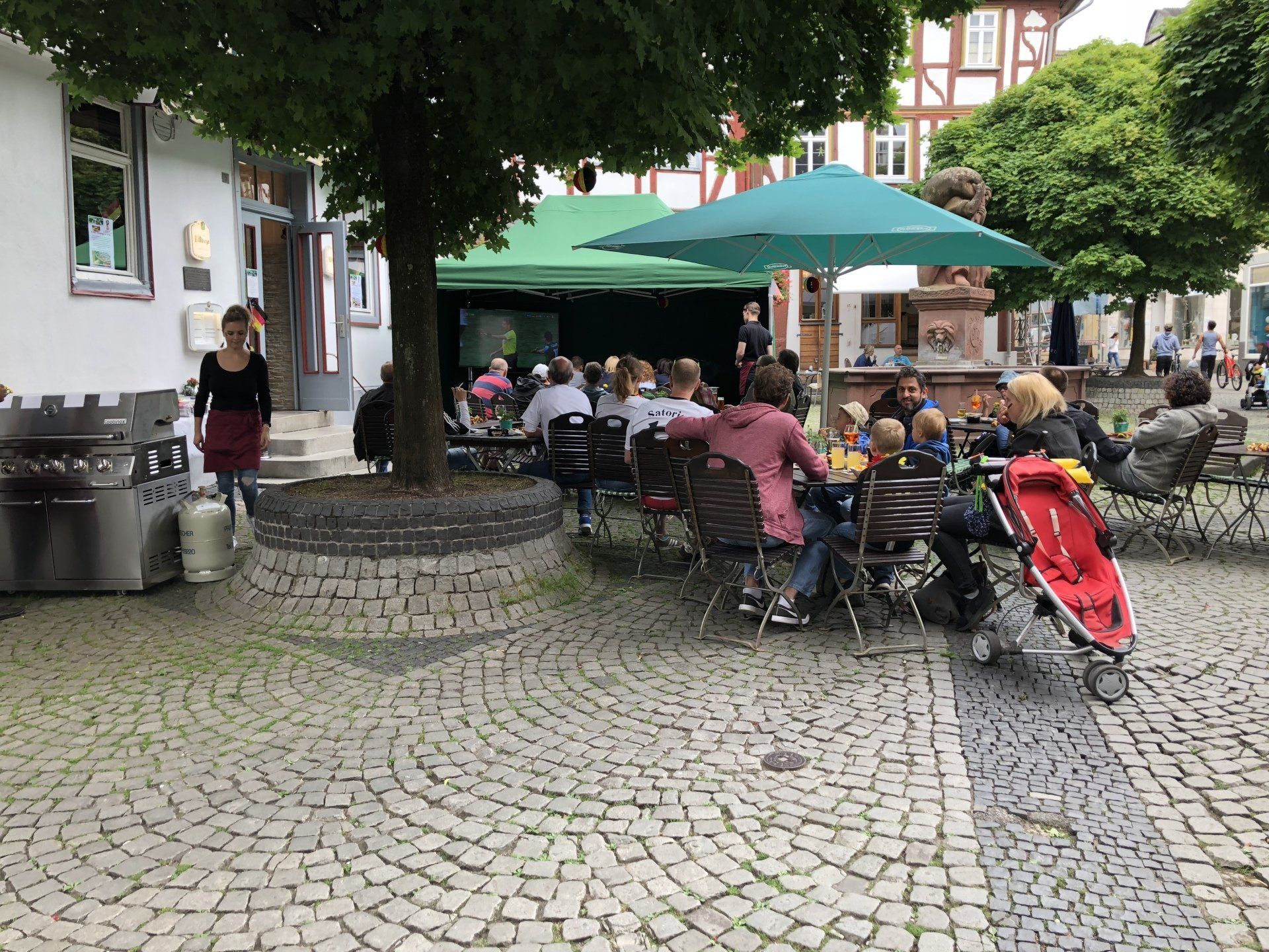 Eine Gruppe von Menschen sitzt auf einem Kopfsteinpflasterplatz an Tischen unter Sonnenschirmen.