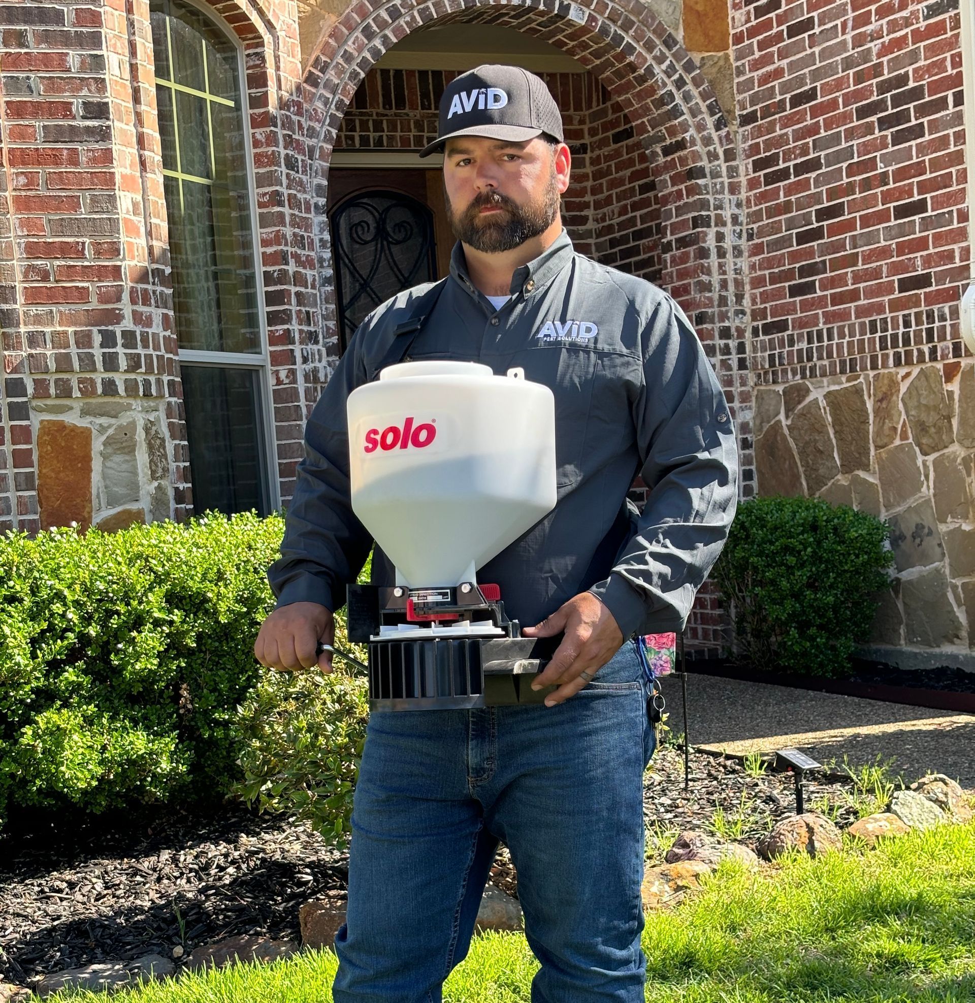 A man is standing in front of a brick house holding a solo spreader.