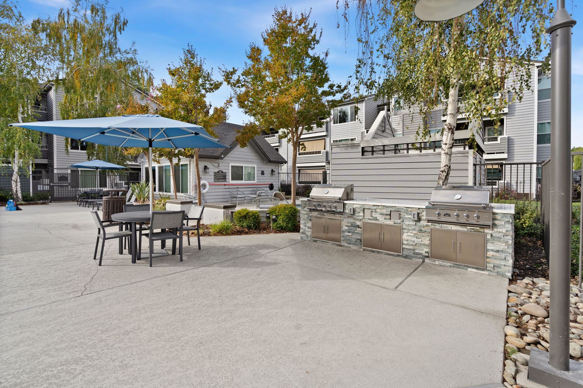 Outdoor community barbecue area with grills, tables, and blue umbrellas.