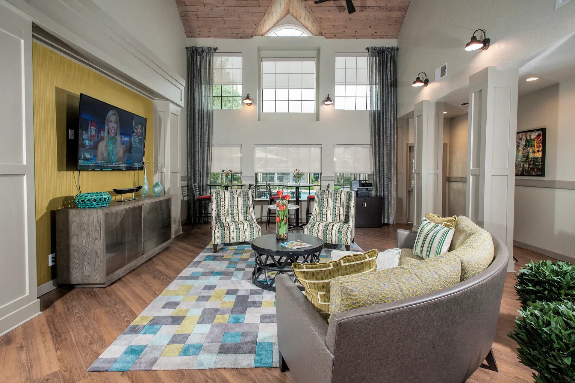 Bright community lounge with sofa, two striped chairs, a wall-mounted TV, and large windows.