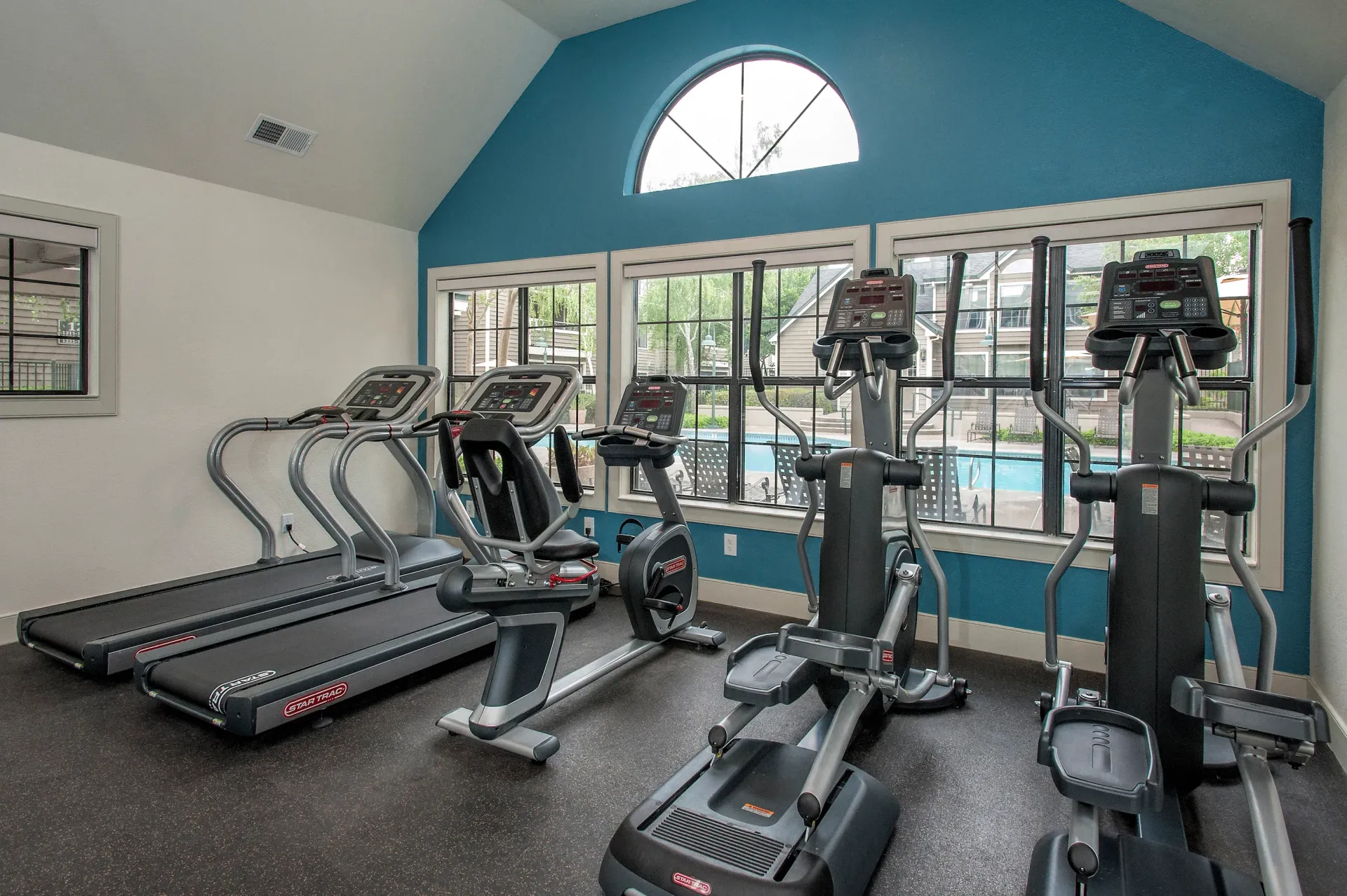 Cardio fitness room with treadmills, ellipticals, and bikes by a large window and blue wall.