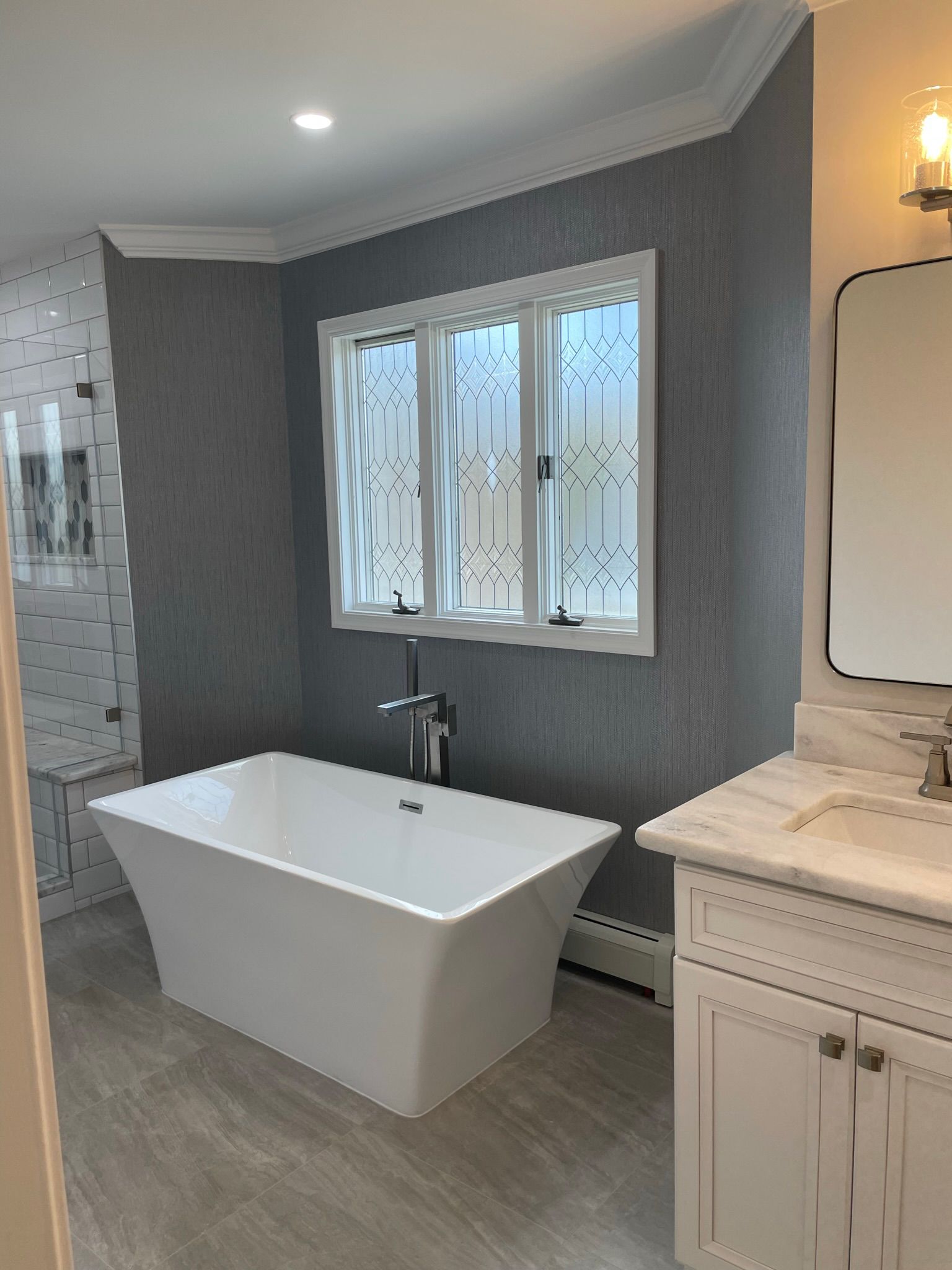 Modern bathroom with freestanding white tub, gray walls, tiled floor, and vanity beside a window Modern bathroom with freestanding white tub, gray walls, tiled floor, and vanity beside a window